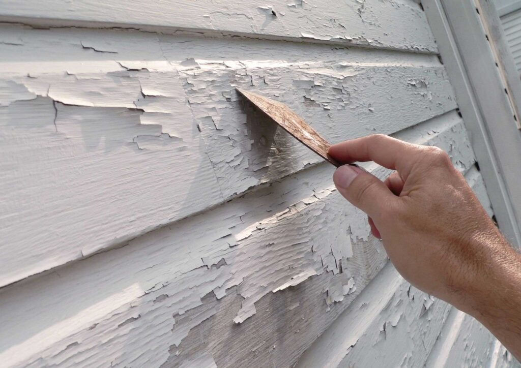 Preparing house exterior for painting by scraping loose paint and sanding siding